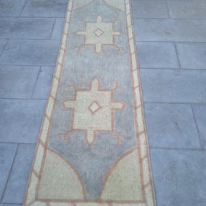 May include: A long, rectangular rug with a light blue background and a beige pattern. The pattern features two large, stylized diamond shapes with smaller diamond shapes inside. The rug has a fringe border.