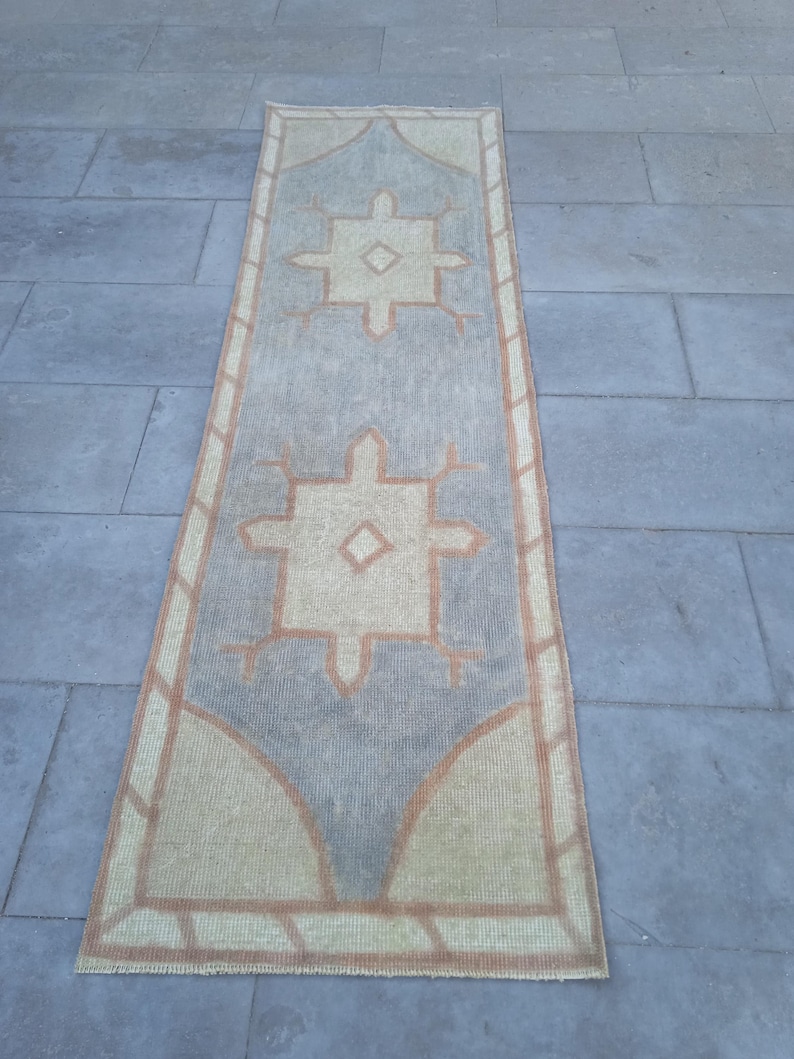 May include: A long, rectangular rug with a light blue background and a beige pattern. The pattern features two large, stylized diamond shapes with smaller diamond shapes inside. The rug has a fringe border.