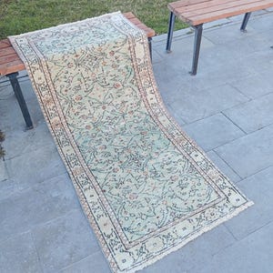 May include: A rectangular, vintage-style rug with a faded floral pattern in shades of green, beige, and rust. The rug is placed on a wooden bench, showcasing its intricate design and fringed edges. The rug's dimensions are not visible.