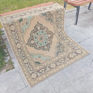 May include: A beige and brown patterned rug with a central floral design in teal and brown. The rug has a border with repeating geometric patterns. The rug is partially draped over a wooden bench, showcasing its design and texture.