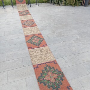 Könnte beinhalten: Ein langer, rechteckiger Läuferteppich mit einem sich wiederholenden Muster aus geometrischen Formen und floralen Motiven. Der Teppich hat einen ziegelroten Hintergrund mit Akzenten in Grün, Blau und Beige. Der Teppich liegt auf einer grauen Steinoberfläche.