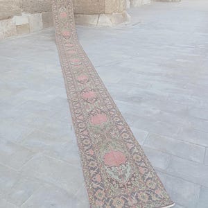 May include: A long, narrow runner rug featuring a repeating floral pattern in shades of dusty pink, sage green, and charcoal gray. The rug has a detailed border and a fringed edge, extending from a vertical position down onto a flat surface.