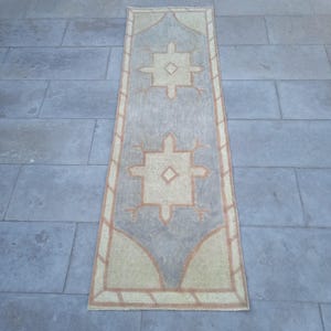 May include: A long, rectangular rug with a beige background and a light gray pattern. The pattern features two large, symmetrical designs with a diamond shape in the center. The rug has a brown border.