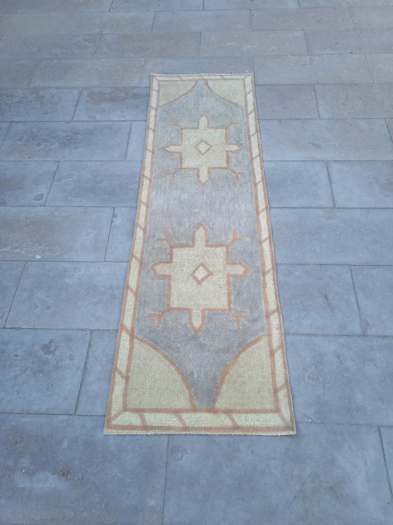 May include: A long, rectangular rug with a beige background and a light gray pattern. The pattern features two large, symmetrical designs with a diamond shape in the center. The rug has a brown border.