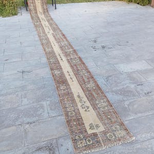 May include: A long, rectangular runner rug with a central beige stripe and patterned borders in shades of brown, red, and green. The rug is placed on a grey stone surface, with a small wooden bench visible in the background.