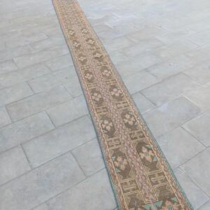 May include: A long, narrow rug with a detailed geometric pattern. The rug features a central design in shades of brown, pink, and teal, with a border in similar colours. The rug is likely made of woven material and is suitable for a hallway or entryway.