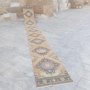 May include: A long, narrow runner rug featuring a repeating pattern of large, ornate floral medallions in shades of muted orange, sage green, and charcoal grey. The rug has a decorative border at the end with small floral motifs and a fringed edge.