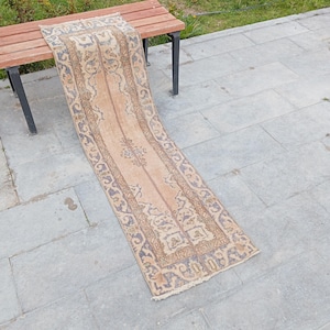 May include: A rectangular, vintage-style rug with a central peach-colored field and intricate border designs in shades of blue and brown. The rug is placed on a wooden bench and gray stone surface, showcasing its detailed patterns and aged appearance.