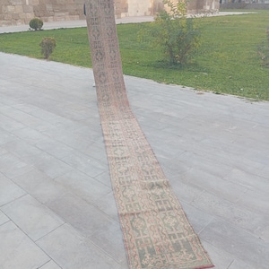 May include: A long, narrow rug with a repeating geometric pattern in shades of green, red, and beige. The rug is rolled out on a gray stone surface, with a building and greenery in the background. The rug's design is visible.