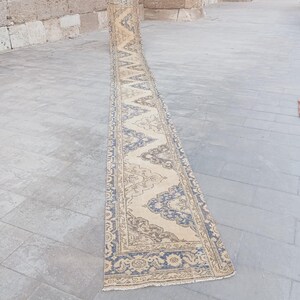 May include: A long, narrow runner rug featuring a repeating geometric pattern in muted blue and beige tones. The design includes intricate floral-inspired motifs within a central zigzag border, finished with short fringe at the ends.