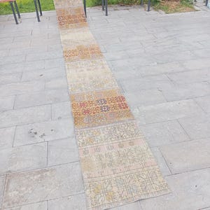 May include: A long, narrow runner rug featuring a repeating patchwork design in muted tones of beige, tan, mustard yellow, and faded red. The textile displays intricate geometric and floral patterns across its entire length, with a weathered appearance.