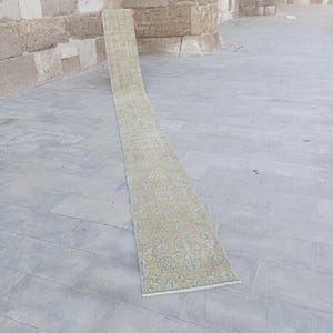 May include: A long, narrow runner rug featuring a muted mustard yellow base color with a repeating light blue floral and vine pattern. The rug has a rectangular shape with short white fringe detailing at the bottom edge and a flat-woven texture.