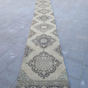 May include: A long, beige runner rug with a brown geometric pattern. The rug has a fringed border and is made of a soft, woven material.