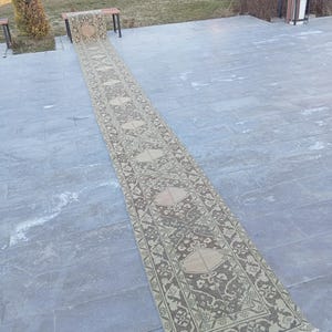 Peut inclure: Un long tapis de couloir rectangulaire avec un motif détaillé en beige et vert olive. Le tapis présente un motif central avec des éléments floraux et géométriques, et une bordure à franges. Le tapis est exposé à l'extérieur.