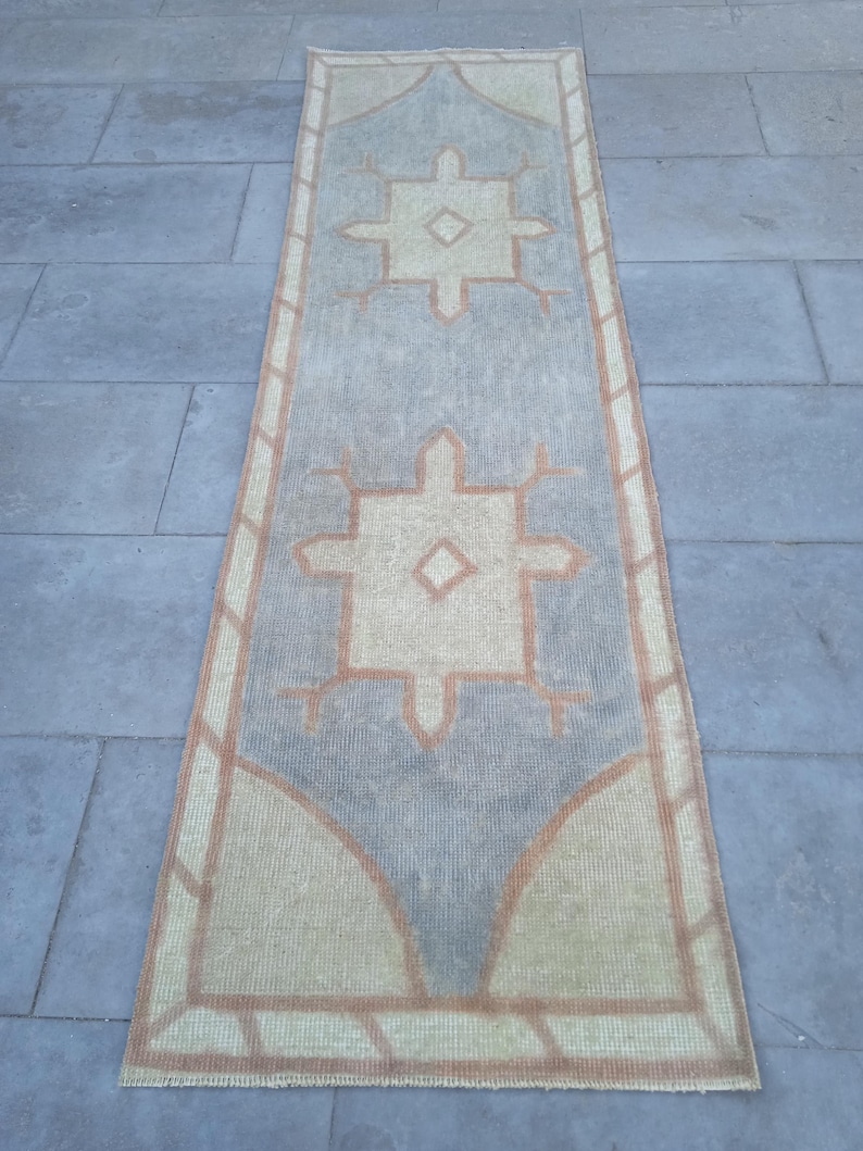 May include: A long, rectangular rug with a geometric pattern in shades of beige, brown, and gray. The rug has a fringed edge and features a repeating design of stylized stars.