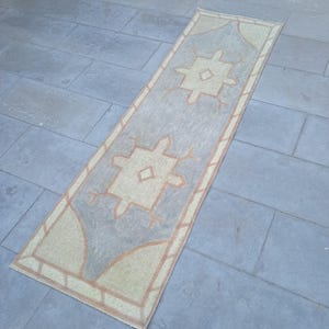 May include: A long, rectangular rug with a light blue background and a beige pattern. The pattern features two large, stylized diamond shapes with smaller diamond shapes inside. The rug has a fringe border.