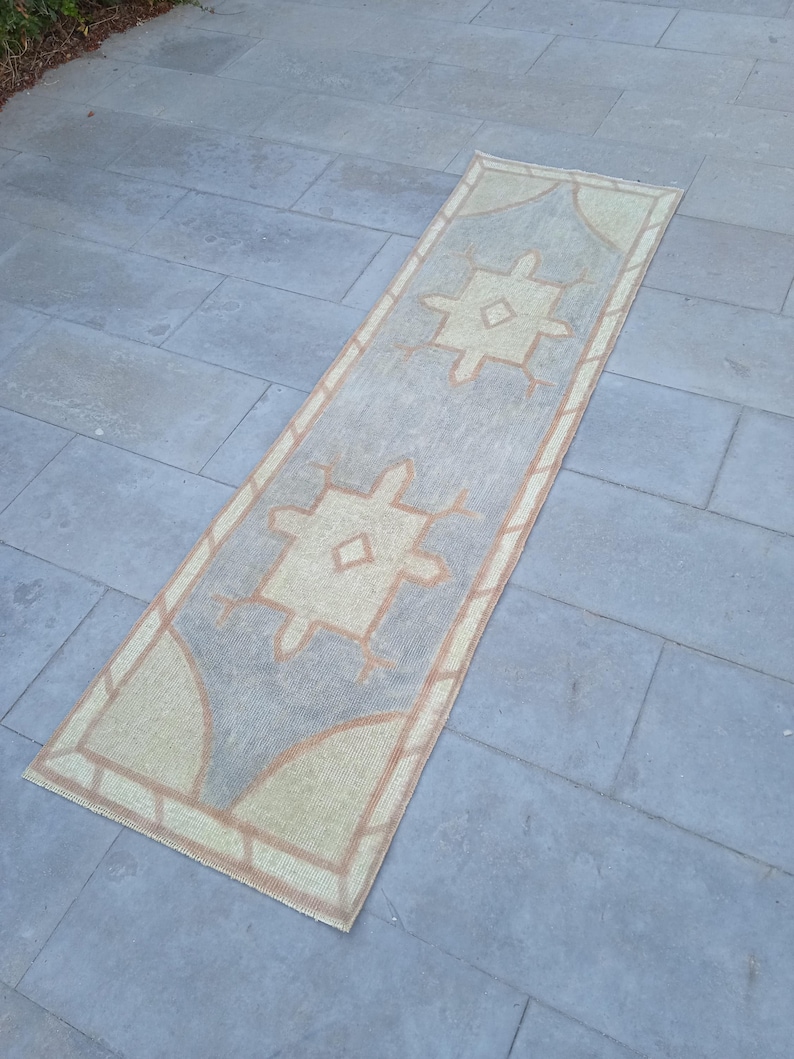 May include: A long, rectangular rug with a light blue background and a beige pattern. The pattern features two large, stylized diamond shapes with smaller diamond shapes inside. The rug has a fringe border.