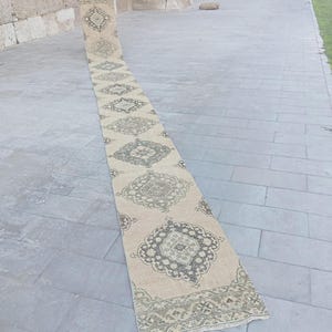 May include: A long, narrow runner rug featuring a beige base colour with a repeating pattern of dark grey, ornate, geometric medallions. The rug has a decorative border at the end and a flat-woven texture, extending across a stone-paved surface.