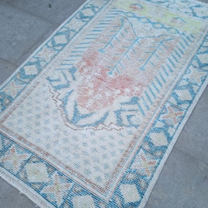 Puede incluir: Una alfombra turca vintage con fondo blanco y un patrón geométrico azul y rosa. La alfombra tiene un borde con flecos.