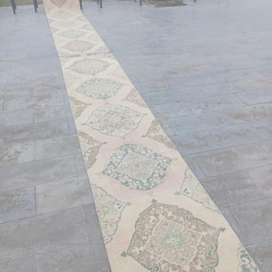 Könnte beinhalten: Ein langer, rechteckiger Teppich mit einem komplizierten, sich wiederholenden Muster aus floralen und geometrischen Designs in Beige-, Rosa-, Grün- und Brauntönen. Der Teppich liegt auf einer grauen Steinoberfläche, mit Bänken und Grün in der Hintergrund.