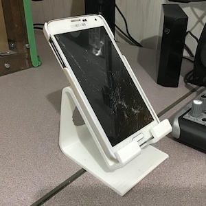May include: A white smartphone with a cracked screen is propped up on a white plastic stand. The stand is angled, with a base and a cradle to hold the phone. The phone is a Samsung model.