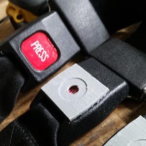 May include: Close-up of black seat belt buckles with a red "PRESS" button. The buckles are made of black plastic and have a silver metal clasp. The seat belts are made of black nylon.