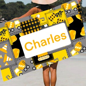 May include: A yellow and gray beach towel with a construction theme. The towel features a variety of construction vehicles, including dump trucks, bulldozers, and cranes. The towel also has a black and white checkered pattern and the name "Charles" printed on it.