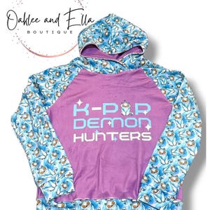 May include: A purple hoodie with blue patterned sleeves and hood. The sleeves and hood feature a repeating pattern of cartoon characters on a blue background. The front of the hoodie has the text "K-POP DEMON HUNTERS" in light blue and white.