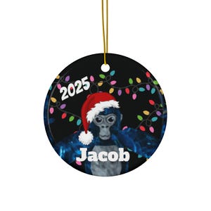 May include: Round Christmas ornament featuring a blue gorilla wearing a Santa hat. The ornament has the year "2025" and the name "Jacob" printed on it. A string of colorful Christmas lights surrounds the gorilla.