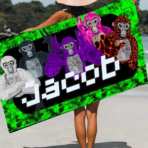 May include: A beach towel with a green and black background featuring five monkeys in different colors and poses. The word "JACO" is written in white letters on the towel.