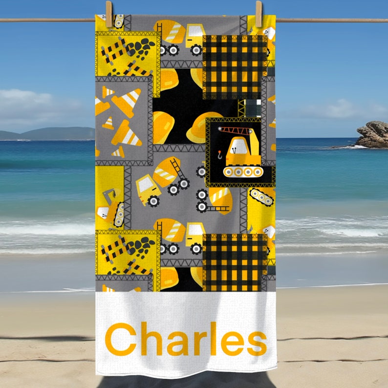 May include: A yellow and grey beach towel with a construction theme. The towel features a repeating pattern of yellow construction vehicles, cones, and hard hats. The towel has a white border with the name "Charles" printed in yellow.