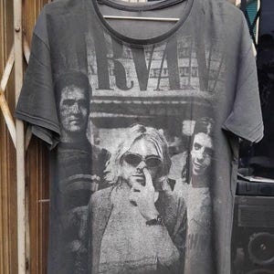 May include: Dark grey t-shirt with a black and white image of the band Nirvana. The band's name is printed in large letters at the top. The image shows three band members, including Kurt Cobain.