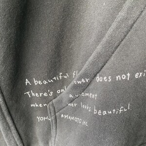 May include: Close-up of a dark gray hoodie with a pocket. White handwritten text is visible on the fabric, including the phrases "A beautiful flower does not exist" and "YOHJI YAMAMOTO inc."
