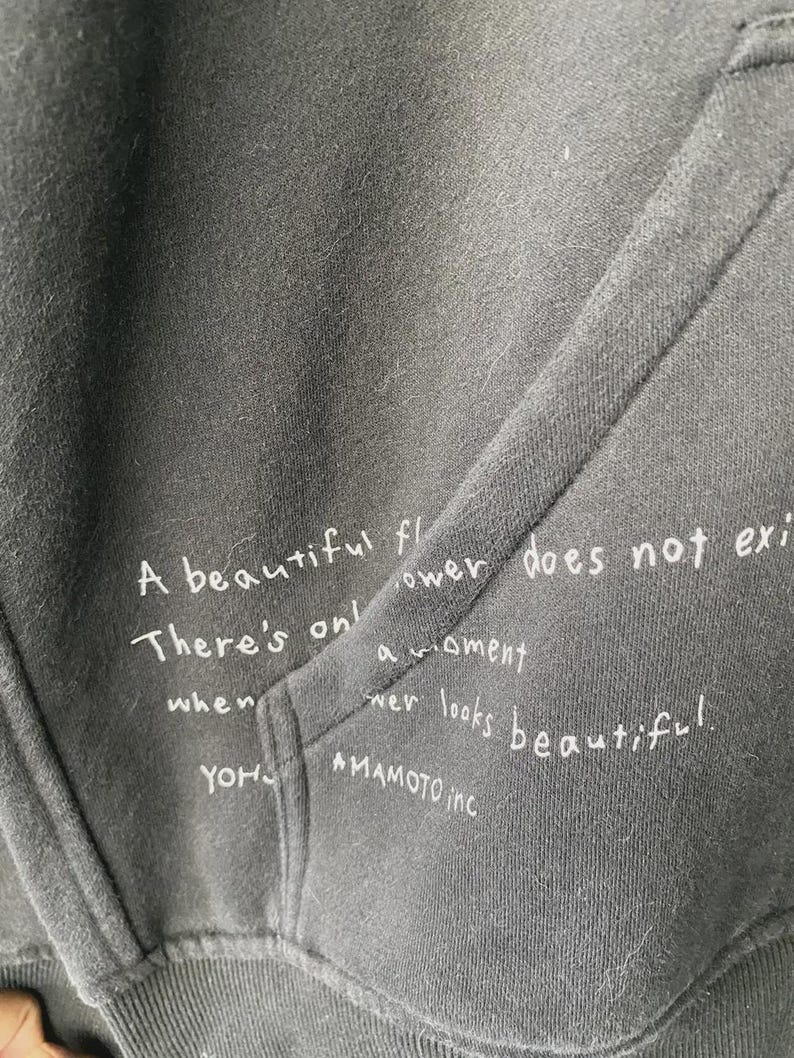 May include: Close-up of a dark gray hoodie with a pocket. White handwritten text is visible on the fabric, including the phrases "A beautiful flower does not exist" and "YOHJI YAMAMOTO inc."