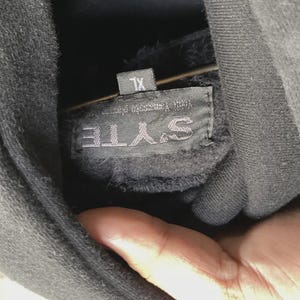May include: Close-up of a black hoodie with a tag that reads "SYTE" and "XL". The tag also has text that is difficult to read. The hoodie is made of a soft, dark material.