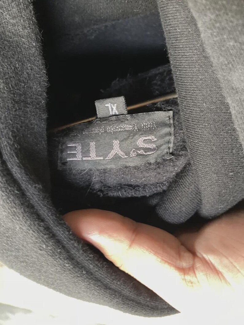 May include: Close-up of a black hoodie with a tag that reads "SYTE" and "XL". The tag also has text that is difficult to read. The hoodie is made of a soft, dark material.