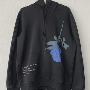 May include: Black hoodie with a blue floral design on the front. The hoodie has a drawstring hood and a front pocket. Text on the front reads: "A beauty never does exist. There's a moment when it is beautiful."