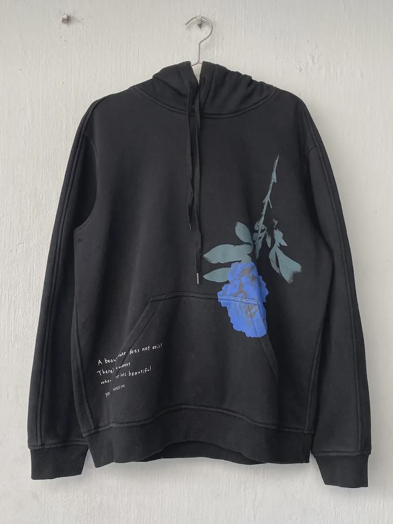 May include: Black hoodie with a blue floral design on the front. The hoodie has a drawstring hood and a front pocket. Text on the front reads: "A beauty never does exist. There's a moment when it is beautiful."