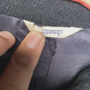 May include: Close-up of a gray fabric garment with a label that reads "dessin" in a light purple font. The label is attached to a dark gray lining. The garment appears to be a coat or jacket.