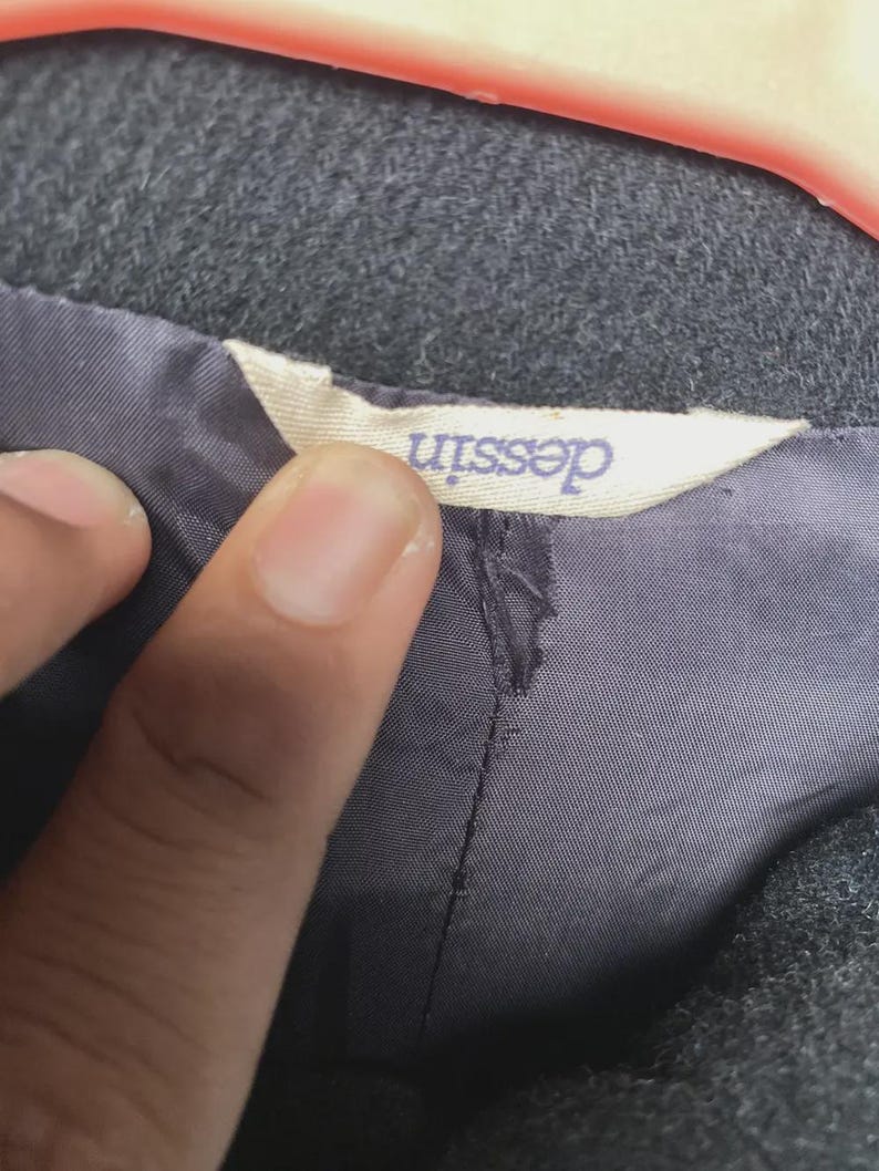 May include: Close-up of a gray fabric garment with a label that reads "dessin" in a light purple font. The label is attached to a dark gray lining. The garment appears to be a coat or jacket.