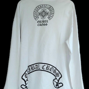 Op de afbeelding: Wit shirt met lange mouwen en zwarte grafische ontwerpen. De achterkant heeft een cirkelvormig ontwerp met de tekst "HEAVEN ORDER BELL 13 BAD LUCK" en "GRIBAL CROSS". Een banner met "GRIBAL CROSS" staat onderaan. Het shirt hangt aan een houten hanger.