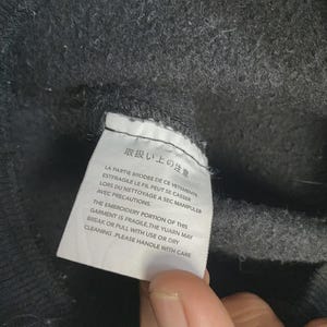 May include: Close-up of a dark gray fabric with a white tag attached. The tag has text in Japanese and English, warning about the fragility of the embroidery and care instructions. The fabric appears to be a sweater or sweatshirt.