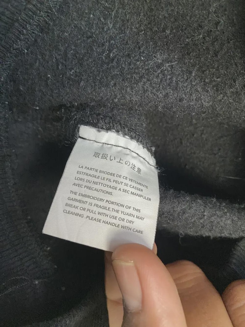 May include: Close-up of a dark gray fabric with a white tag attached. The tag has text in Japanese and English, warning about the fragility of the embroidery and care instructions. The fabric appears to be a sweater or sweatshirt.