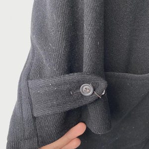 May include: Close-up of a black wool coat sleeve with a buttoned cuff. The cuff is folded back, revealing the interior fabric. The coat appears to be made of a thick, textured material.
