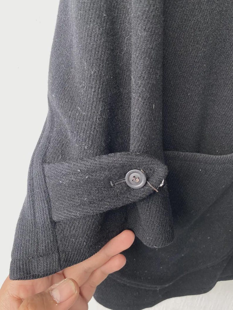 May include: Close-up of a black wool coat sleeve with a buttoned cuff. The cuff is folded back, revealing the interior fabric. The coat appears to be made of a thick, textured material.