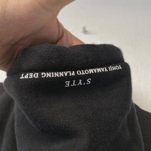 May include: Black sweatshirt collar with white text that reads "S'YTE YOHJI YAMAMOTO PLANNING DEPT". The fabric appears to be a soft, dark material. The image is a close-up of the collar.