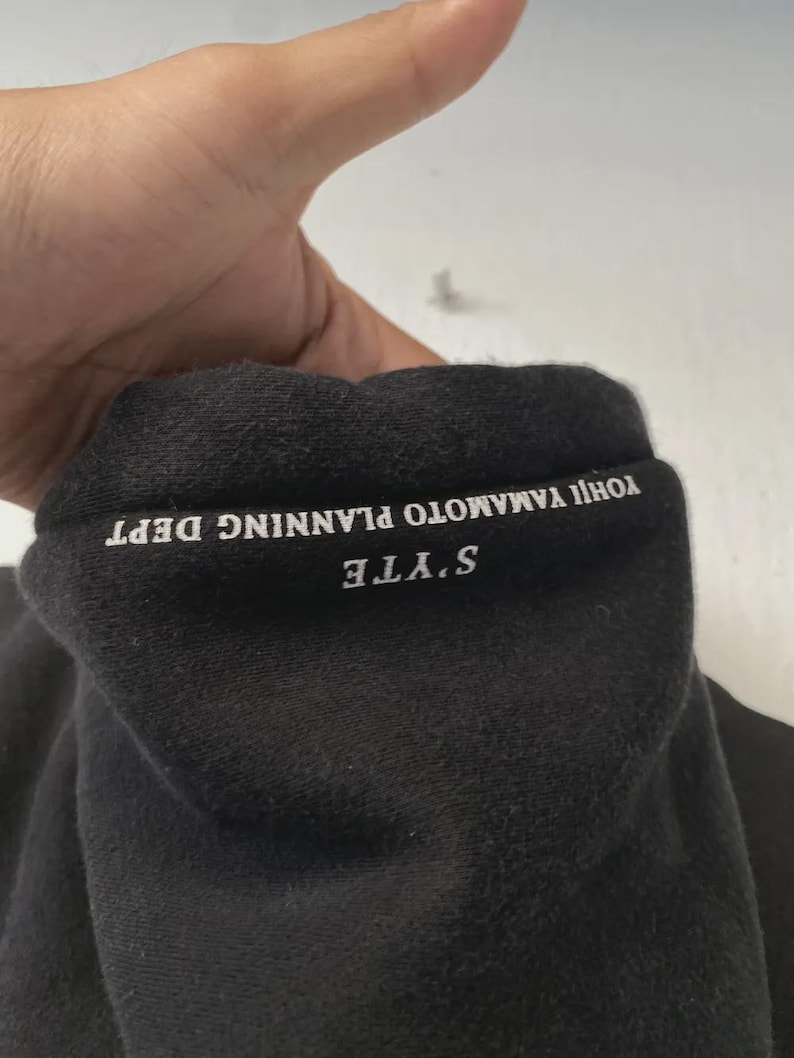 May include: Black sweatshirt collar with white text that reads "S'YTE YOHJI YAMAMOTO PLANNING DEPT". The fabric appears to be a soft, dark material. The image is a close-up of the collar.