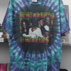 Peut inclure: T-shirt tie-dye avec un graphisme de Ziggy Marley and the Melody Makers. Le t-shirt présente un motif tourbillonnant dans les tons de bleu, violet et vert. Le nom du groupe est imprimé en rouge, jaune et vert.