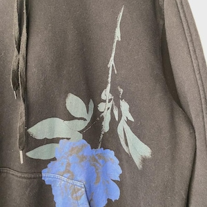 May include: Dark gray hoodie with a large blue and gray floral design on the front. The hoodie has a drawstring hood and a front pocket. The floral design is a stylized rose.