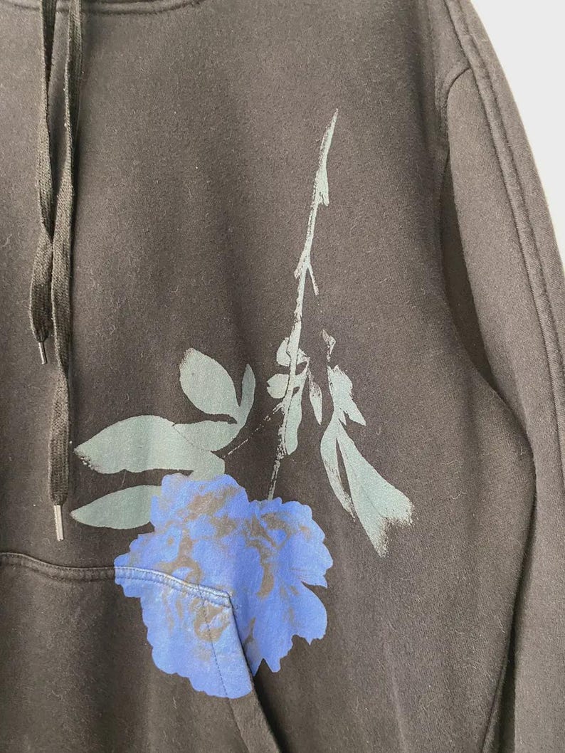 May include: Dark gray hoodie with a large blue and gray floral design on the front. The hoodie has a drawstring hood and a front pocket. The floral design is a stylized rose.